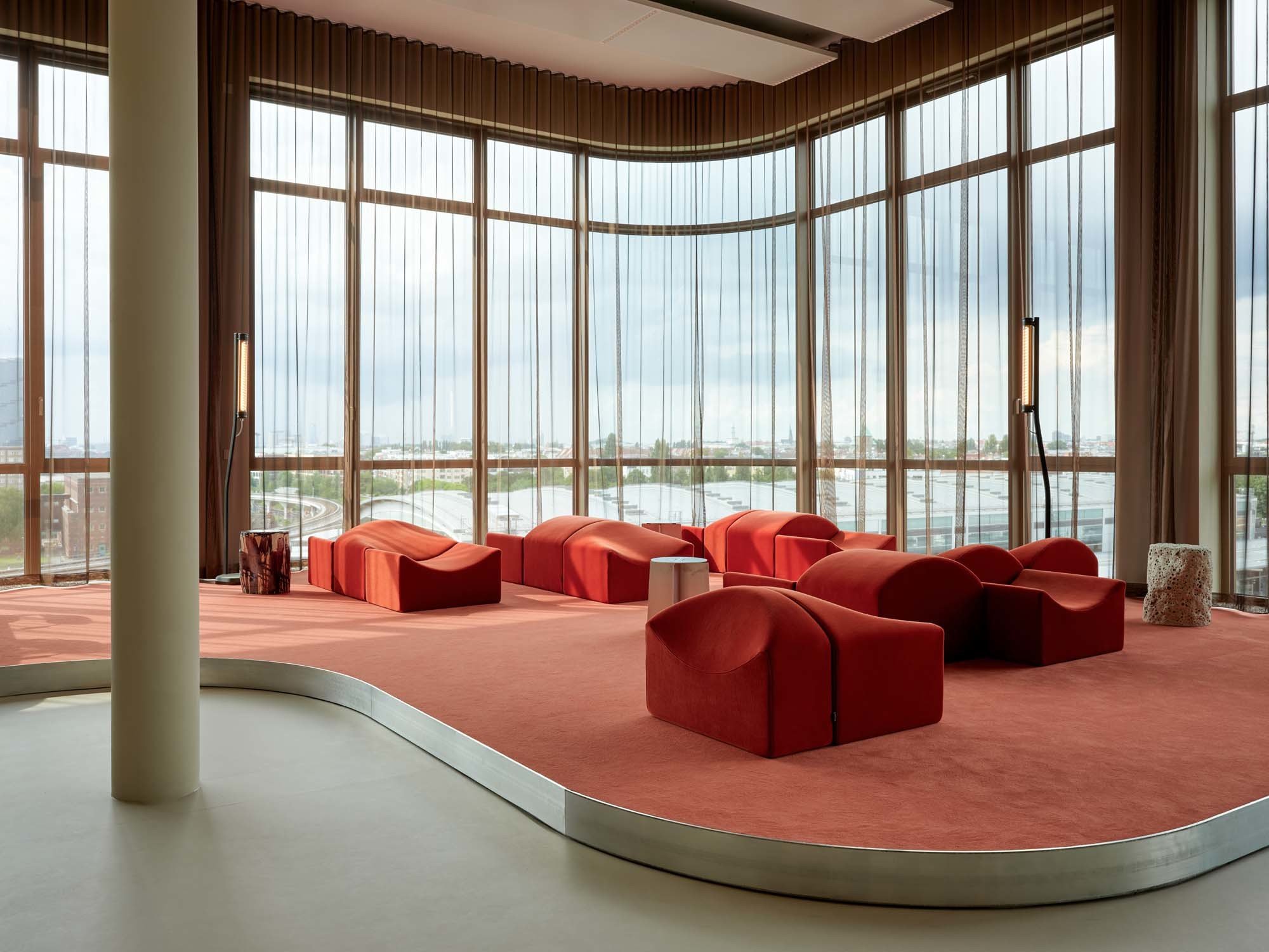 Die Community-Area ist wie eine Bühne gestaltet. Auf dem roten Podest steht das modulare Sofa Asmara von Ligne Roset, das von puristischen Sophie.Stool-Hockern von Loes.Beta flankiert wird.