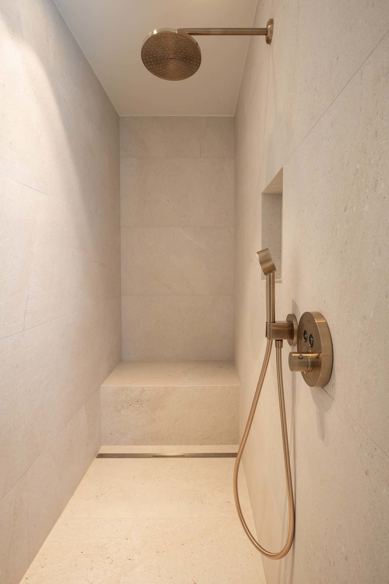 Die schwellenlose Dusche mit Sitzbank wird über das DallFlex-Entwässerungssystem und die Rinne CeraFloor Select in Messing matt von Dallmer entwässert.