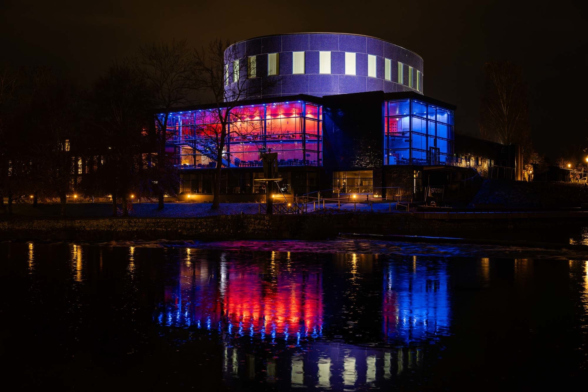 Das Gävle Konserthus bei Nacht, Foyer in rotem und blauem Licht, gespiegelt im Gavleån.